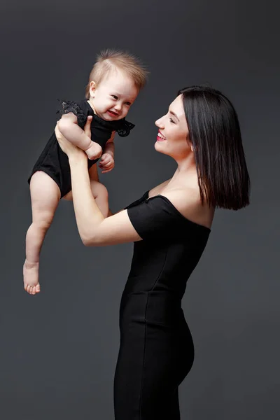 Élégante jeune mère en robe noire tient bébé fille en studio sur fond gris — Photo