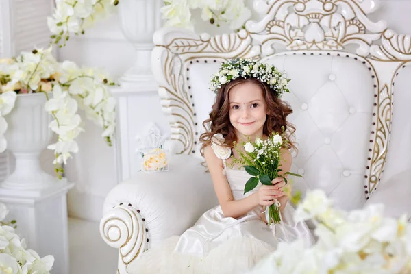 Schattig klein meisje met donker haar in een lichte studio met een krans van bloemen en een boeket. Gelukkig kindertijd concept. — Stockfoto