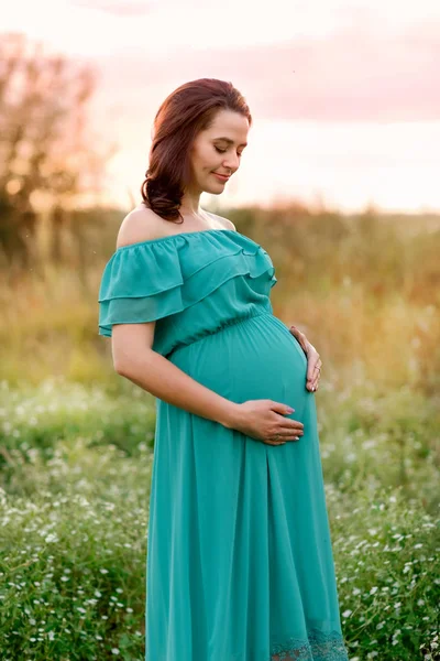 Bella donna incinta in un abito verde si trova nel giardino di salice e guarda la pancia il giorno d'estate — Foto Stock