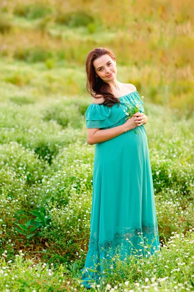 美丽的孕妇穿着绿色礼服站在柳树花园，看着相机在夏天的一天 — 图库照片