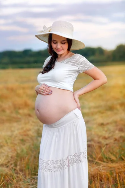 Gadis hamil lucu dalam topi jerami dan rok putih berdiri di lapangan saat matahari terbenam andand melihat perutnya yang besar — Stok Foto
