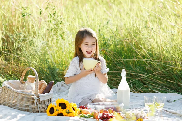 Schattig klein meisje in de witte kleren zittend op het veld drinken van melk en glimlachend. Stockafbeelding