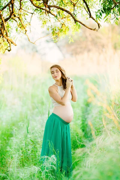 Słodka, uczciwa dziewczyna w ciąży w zielonej spódnicy stoi w parku z jej oczy zamknięte i warkocze jej warkocz. — Zdjęcie stockowe