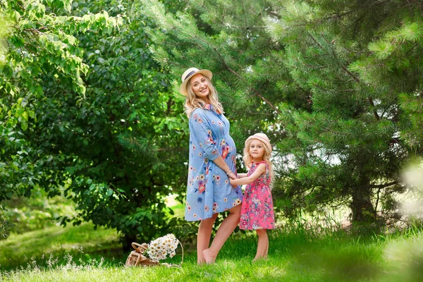 Una hermosa madre embarazada con un sombrero de paja está de pie junto a su hija pequeña en un parque en un día soleado de verano. El concepto de relaciones familiares felices . —  Fotos de Stock