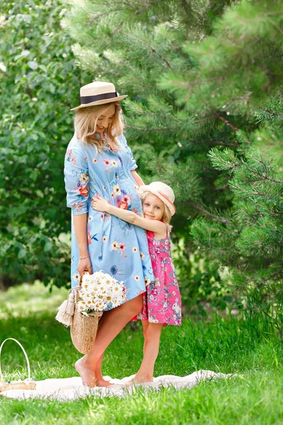 Beautiful pregnant girl in a straw hat is standing with a bouquet of summer flowers next to her little daughter and hugging in the park on a sunny summer day. — Stock Photo, Image