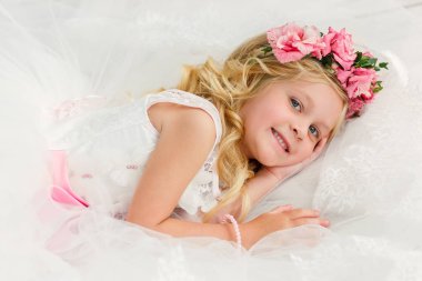 Çiçek bir çelenk ile parlak bir stüdyoda sarı saçlı sevimli küçük kız güzel bir beyaz elbise yatıyor. Mutlu çocukluk konsepti.
