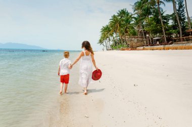 Beyaz elbiseli anne ve beyaz gömlekli küçük oğlu deniz kıyısında yürüyorlar. Geri görünüm