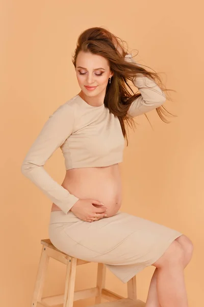 Ciężarna piękna dziewczyna w długim beżu ubrania na beżowym tle w studiu. Modny styl dla kobiet w ciąży. Lampa studyjne. — Zdjęcie stockowe