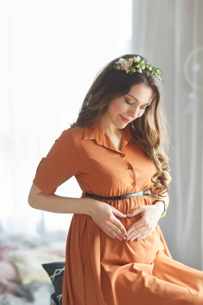 Une belle jeune fille enceinte avec une couronne de fleurs sur la tête dans une robe brune avec de la dentelle est assise près de la fenêtre et tenant ses mains sur le ventre — Photo