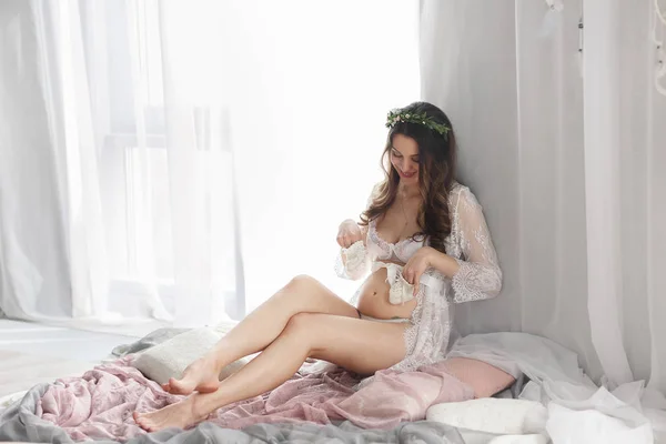 Dantel ile beyaz bir elbise kafasına çiçek çelenk ile güzel bir genç hamile kız pencerenin yanında oturan ve bebek booties elinde tutarak — Stok fotoğraf