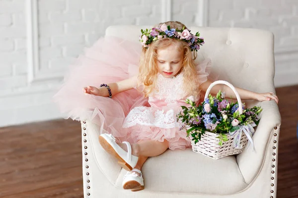 A cute kid - girl in a in a pink dress and flower crown. Beautiful little girl in a bright studio sits, lauht and looks out the window and holding a basket of fresh flowers. — Stock Photo, Image