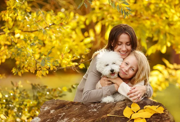Feliz familia mamá e hija con pequeño abrazo de perro blanco y risa soleado día de otoño en el fondo de hojas amarillas —  Fotos de Stock