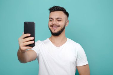 Yakışıklı genç sakallı hippi telefon kullanarak ve parlak bir gülümseme ile selfie alarak beyaz tişört içinde.