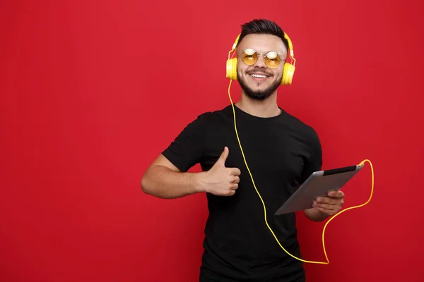 Young bebaarde man in gele zonnebril en hoofdtelefoon poseren met tablet en het tonen van een duim omhoog gebaar op rode achtergrond weg wijzen. — Stockfoto
