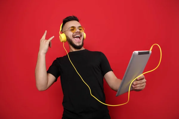 Luminoso uomo barbuto in occhiali da sole gialli e t-shirt nera ascoltare musica sul tablet con volto felice su sfondo rosso . — Foto Stock