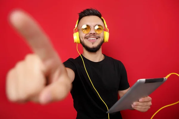 Giovane uomo barbuto in occhiali da sole gialli e cuffie posa allegramente con tablet e su sfondo rosso che punta lontano . — Foto Stock