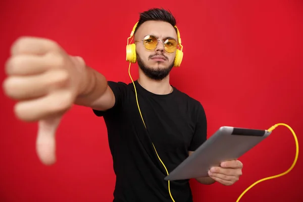 Молодой бородатый мужчина в желтых солнцезащитных очках и наушниках позирует с планшетом и показывает жест вниз на красном фоне, указывая на . — стоковое фото