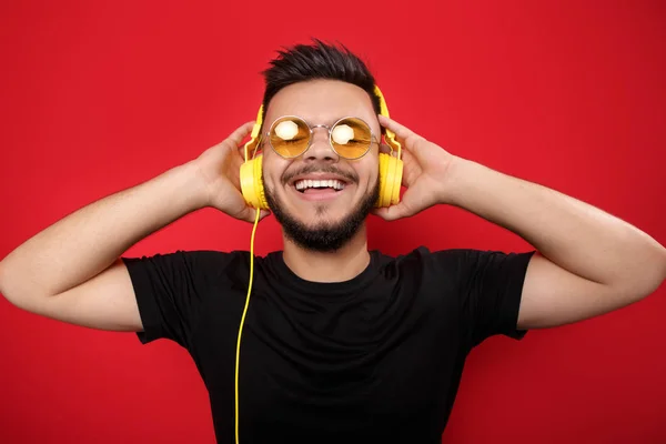 Vrolijke bebaarde jongeman dragen van een gele zonnebril luisteren naar muziek met gele koptelefoon op rode achtergrond. — Stockfoto