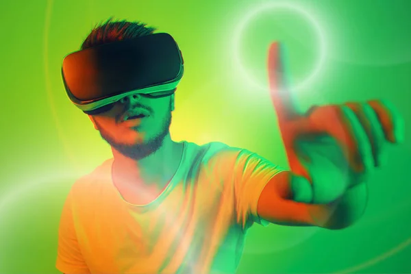 Attractive bearded man trying VR headset and poniting in lightning circle. Young man exploring another world with virtual reality goggles on the colorful background.