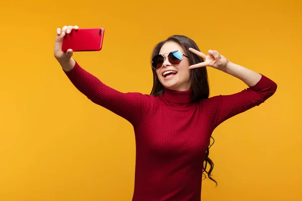 Attractive young lady in stylish sunglasses taking selfie posing in studio in front of orange background. — Stock Photo, Image