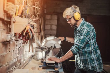 Mesleği marangoz Oluşturucusu testere ile bir daire testere atölyede ahşap bir masa ahşap bir gemide tarafından kulak koruyucular adamla genç hipster sakallı.