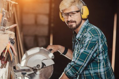 Mesleği marangoz Oluşturucusu testere ile bir daire testere atölyede ahşap bir masa ahşap bir gemide tarafından kulak koruyucular adamla genç hipster sakallı.