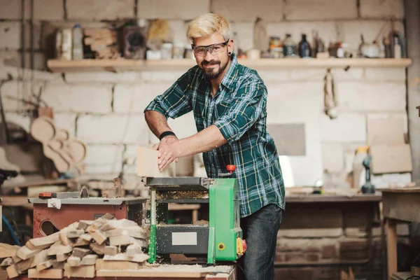 Attraktiver junger Mann mit Hipster-Bart und Augenschutz von Beruf Zimmerermeister sägt mit der Kreissäge ein Holzbrett auf einem Holztisch in der Werkstatt. — Stockfoto