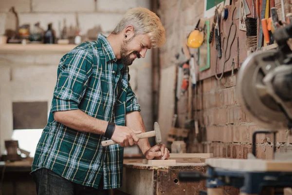 Attraktive blonde junge Hipster bärtiger Mann von Beruf Schreiner Baumeister nagelt Holzbretter mit Hammer auf einem Holztisch in der Werkstatt. — Stockfoto