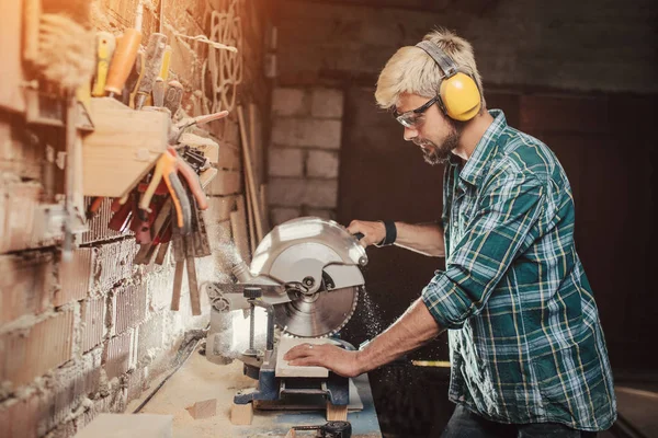 Junger Hipster bärtiger Mann mit Ohrenschutz von Beruf Schreiner Baumeister sägt mit der Kreissäge ein Holzbrett auf einem Holztisch in der Werkstatt. — Stockfoto
