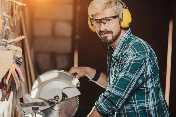 Junger Hipster bärtiger Mann mit Ohrenschutz von Beruf Schreiner Baumeister sägt mit der Kreissäge ein Holzbrett auf einem Holztisch in der Werkstatt. — Stockfoto