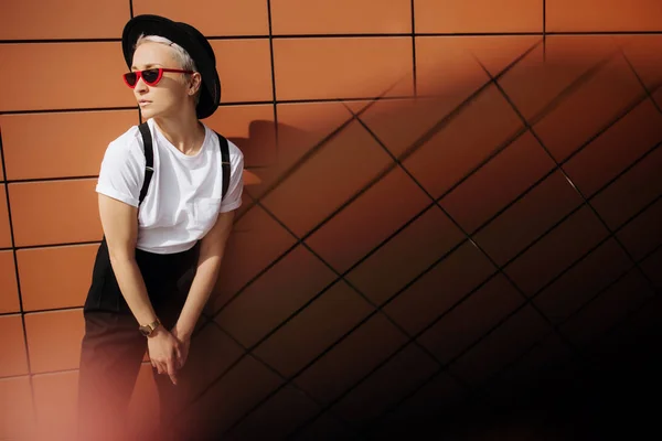 Atraktivní módní hipster mladá žena s stylové červené brýle a černý klobouk zůstat blízko zdi. — Stock fotografie