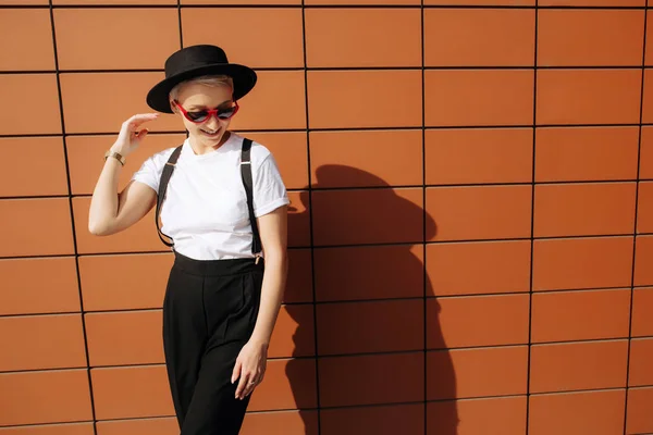 Atractiva mujer joven hipster de moda con gafas de sol rojas con estilo y sombrero negro permanecer cerca de la pared . — Foto de Stock