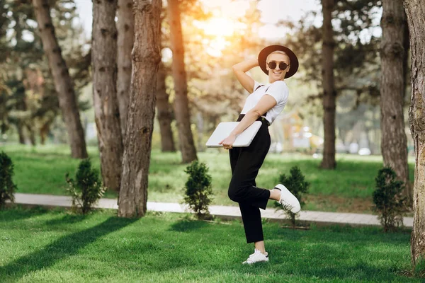 Attraktive trendige fröhliche Hipster junge Frau mit Notizbuch in der einen Hand, Hut und Sonnenbrille haben Spaß im Park bei Sonnenuntergang — Stockfoto