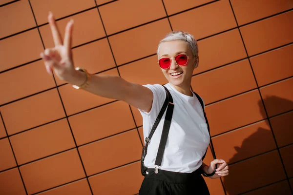 Attraktiva trendiga hipster ung kvinna vistelse nära väggen poserar visar fred hand tecken. — Stockfoto
