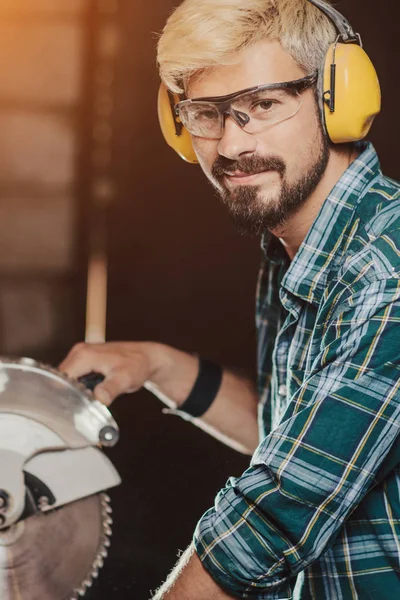 Heller junger Mann mit Hipster-Bart und Ohrenschutz von Beruf Zimmerermeister sägt mit der Kreissäge ein Holzbrett auf einem Holztisch in der Werkstatt. — Stockfoto