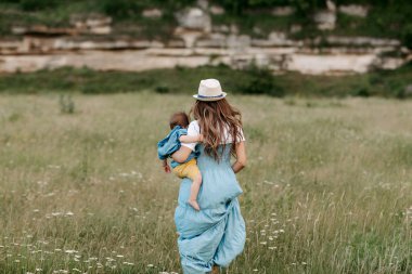 Mutlu genç anne littlte bebek oğlu ile çiçekli dağ yoluyla alan üzerinde yürüyen uzun mavi bir elbise içinde. Arkadan görünüm.