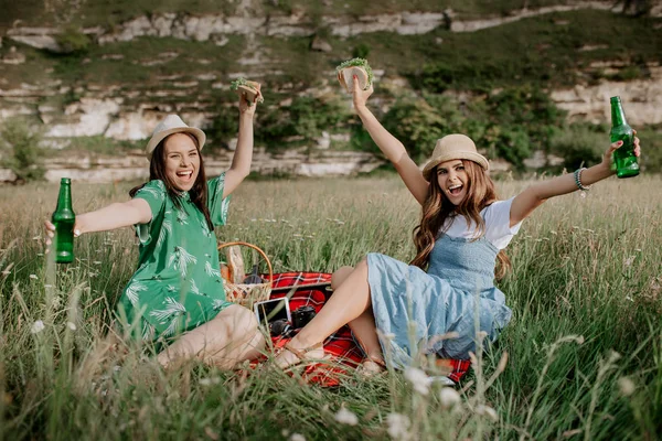İki çekici genç kadın oturup piknik battaniye var aşağı ve eğlenin. Bira ve sandviç. — Stok fotoğraf