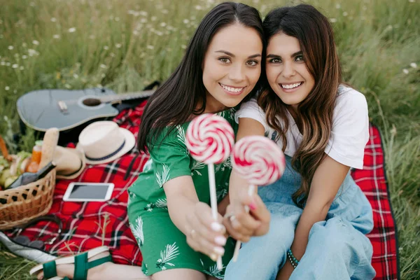 Dua wanita muda cantik dengan senyum indah duduk di atas selimut piknik dengan permen . — Stok Foto