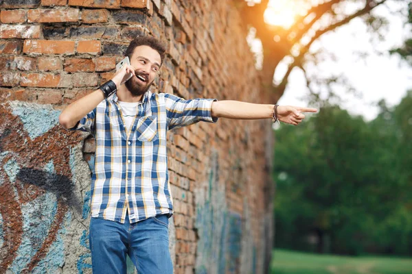 Aantrekkelijke jonge bebaarde hipster met mobiele telefoon wijzen gelukkig weg. — Stockfoto