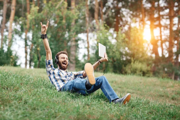 Çimenlerin üzerinde oturan gülümseyen Genç sakallı hippi. Sakallı adam tablet ve parkta müzik dinlerken kulaklık.
