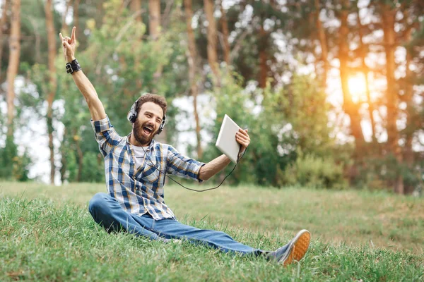 Çimenlerin üzerinde oturan gülümseyen Genç sakallı hippi. Sakallı adam tablet ve parkta müzik dinlerken kulaklık.