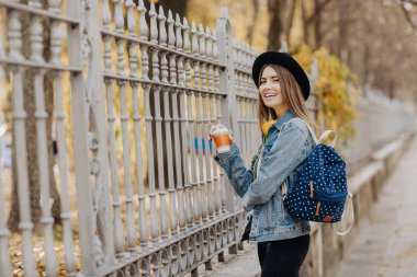 Kahverengi saçlı bir şapka ve fincan çay ya da kahve parkta tutan sırt çantası takmış parlak gülümseyen bir hipster kız resim