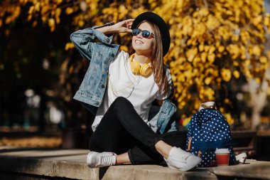 Bir şapka, güneş gözlüğü ve fincan çay ya da kahve parkta tutan sırt çantası kahverengi saçlı parlak gülümseyen bir hipster kız resim