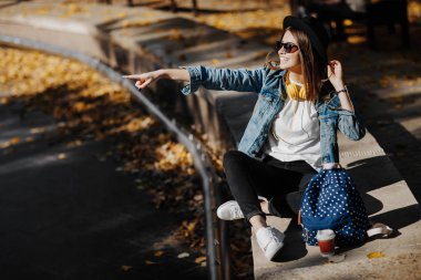 Bir şapka, güneş gözlüğü ve fincan çay ya da kahve parkta tutan sırt çantası kahverengi saçlı parlak gülümseyen bir hipster kız resim