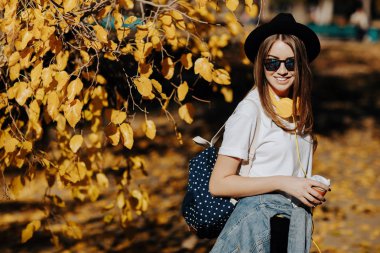 Bir şapka, güneş gözlüğü ve fincan çay ya da kahve parkta tutan sırt çantası kahverengi saçlı parlak gülümseyen bir hipster kız resim.