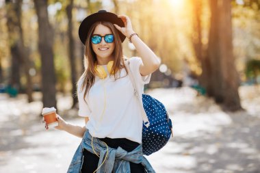 Bir şapka, güneş gözlüğü ve fincan çay ya da kahve parkta tutan sırt çantası kahverengi saçlı parlak gülümseyen bir hipster kız resim