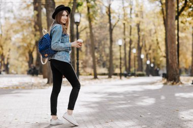 Bir şapka, güneş gözlüğü ve fincan çay ya da kahve parkta tutan sırt çantası kahverengi saçlı parlak gülümseyen bir hipster kız resim