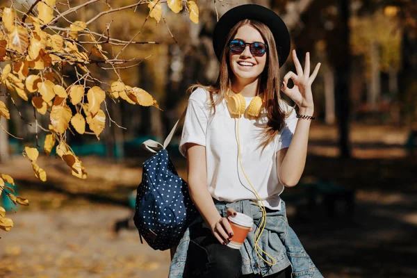 Imagen de una chica hipster sonriente brillante con cabello castaño usando un sombrero, gafas de sol y mochila sosteniendo una taza de té o café en el parque — Foto de Stock