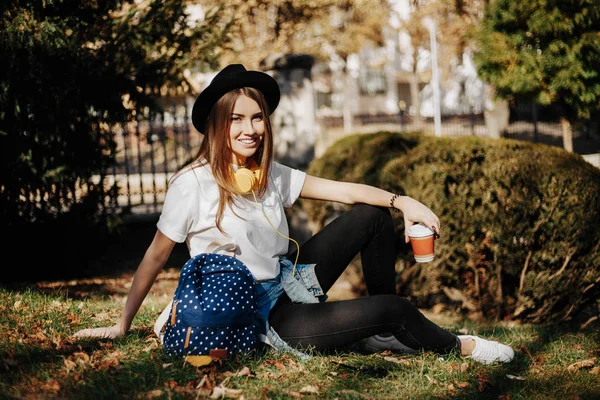 Bild eines strahlend lächelnden Hipstermädchens mit braunen Haaren, das einen Hut, Sonnenbrille und Rucksack mit einer Tasse Tee oder Kaffee in der Hand trägt. junge Frau sitzt auf dem Gras im Park — Stockfoto