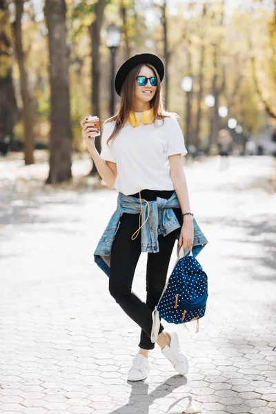 Bir şapka, güneş gözlüğü ve fincan çay ya da kahve parkta tutan sırt çantası kahverengi saçlı parlak gülümseyen bir hipster kız resim — Stok fotoğraf